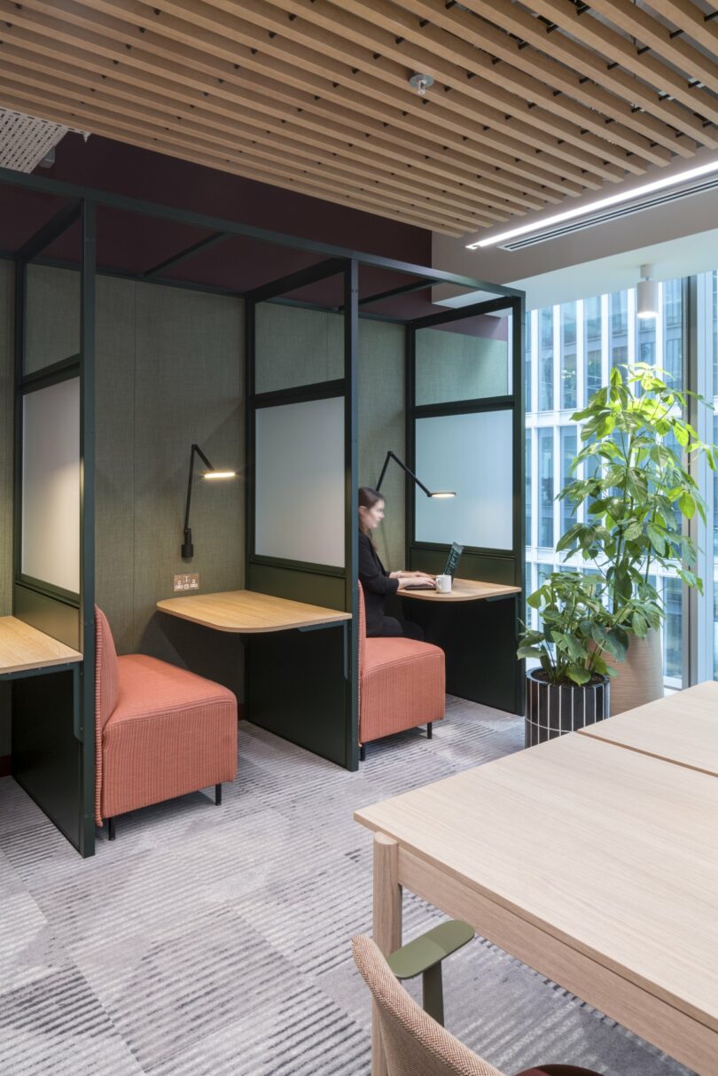 A person works on a Virgin 02 laptop in a modern office cubicle with a wooden ceiling, partitioned seating, and a potted plant nearby.