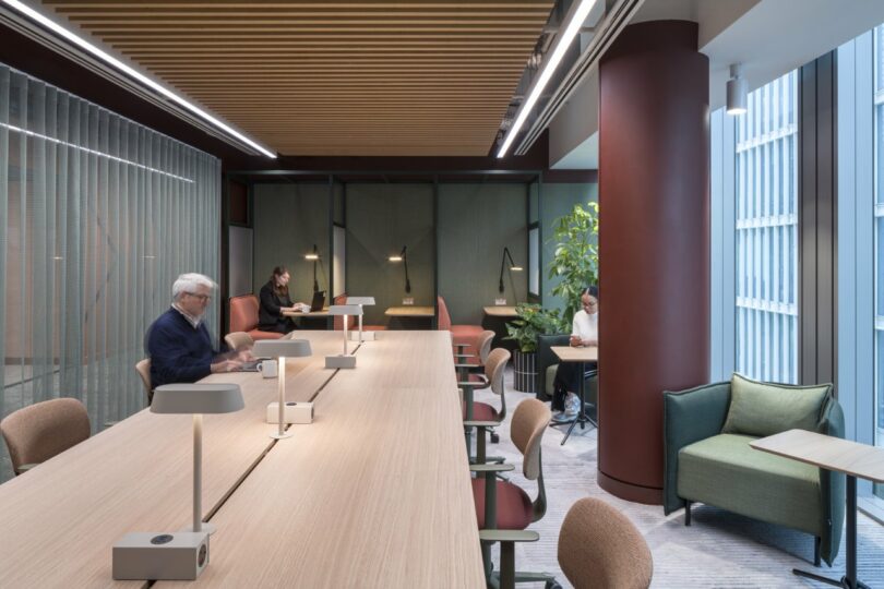 In a modern Virgin 02 office space, a long wooden table is surrounded by chairs and lamps. Three people are seated, absorbed in work or reading. Large windows and plants infuse the room with natural light and greenery.