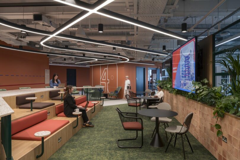 Modern Virgin office space with tiered seating, tables, and chairs. A large screen displays an image as people engage in conversation. Green plants decorate the area, creating a refreshing ambiance akin to O2's signature style.