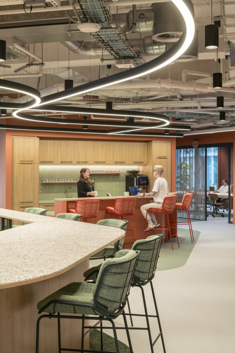 Two people converse at the Virgin 02 kitchen counter in a modern office space with red chairs, curved lighting, and open ceilings.