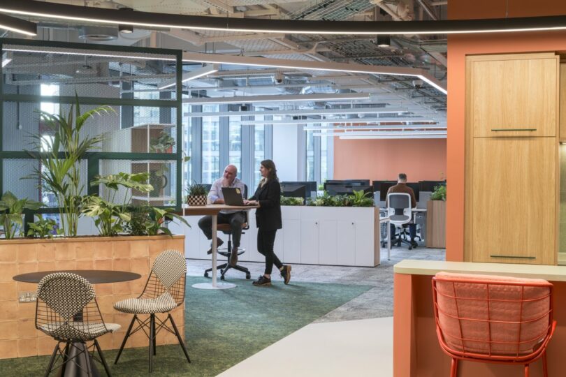 Open office space with modern Virgin 02 furniture. Two people are working on a laptop at a sleek standing desk. Green plants and natural light create a bright, invigorating atmosphere.