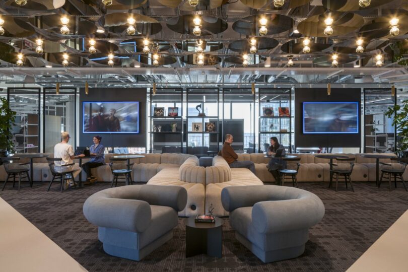 The Virgin 02 modern office lounge features curved sofas, sleek overhead lighting fixtures, and wall-mounted screens. People are seated and conversing in groups.