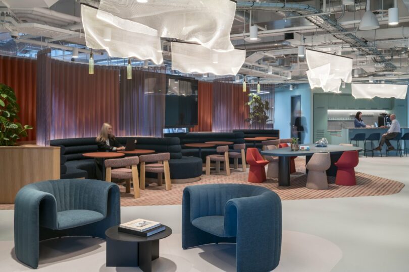 The modern Virgin 02 office lounge features blue chairs and round tables beneath cloud-like light fixtures. People chat in a relaxed atmosphere, while a sleek bar area provides the perfect backdrop.