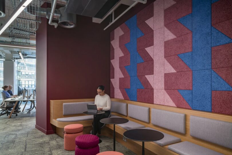 A person sits on a cushioned bench in a Virgin 02 office, using a laptop amidst sleek geometric patterned wall panels. Tables and stools are in the foreground while others work diligently in the background, creating an atmosphere of focused productivity.