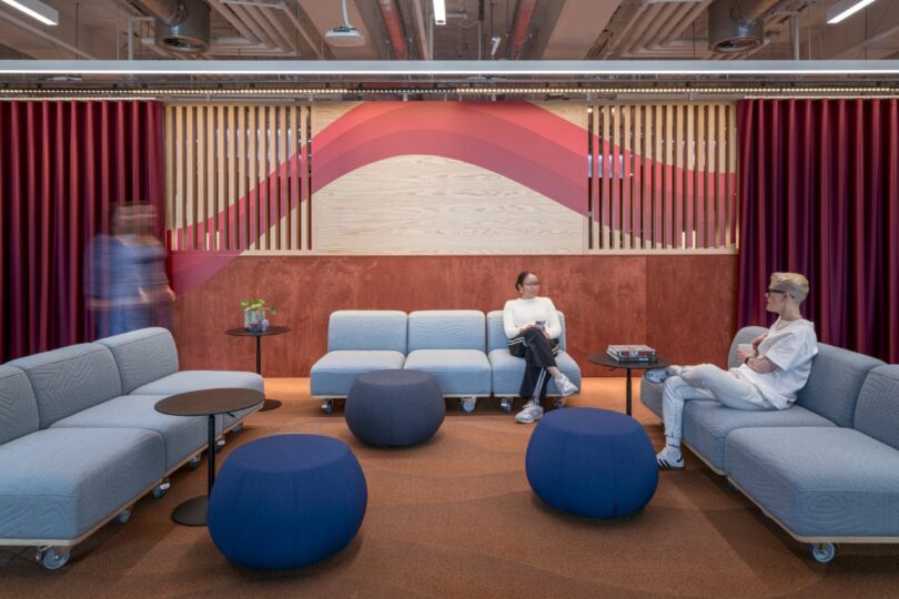 The Virgin 02 modern lounge features blue couches and round ottomans. Two people are seated, conversing against a striking red and wooden wall with an abstract pattern. Another person walks by, immersed in their own world.