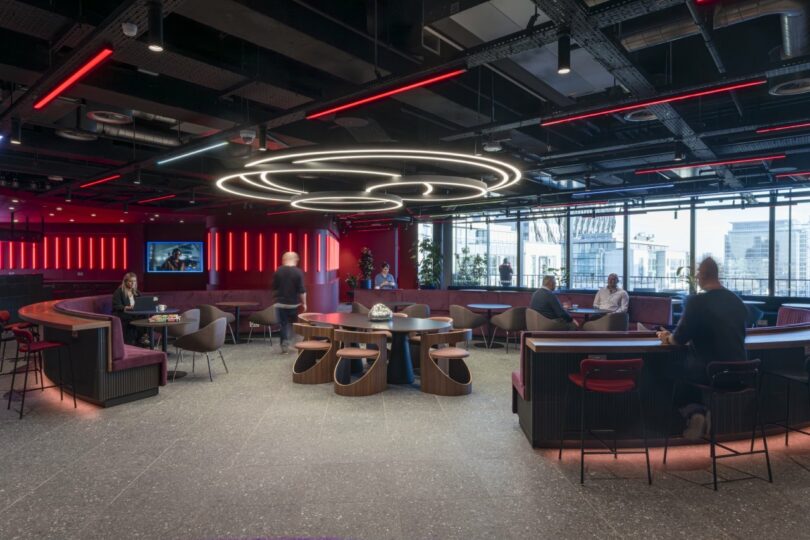 The Virgin 02 lounge area boasts modern circular seating, illuminated by sleek overhead ring lights and surrounded by floor-to-ceiling windows. People relax and stroll amidst a striking red and black color scheme, creating an inviting yet dynamic atmosphere.