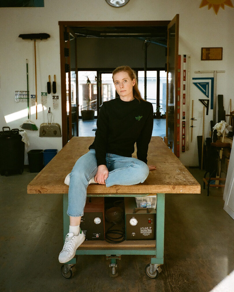 Person sitting on a wooden workbench in a workshop, wearing a black sweater and jeans. Various tools and equipment are visible in the background