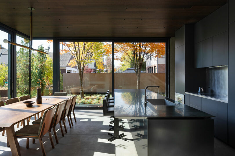 Modern kitchen with large windows, black island countertop, wooden dining table, and views of trees and neighboring houses.