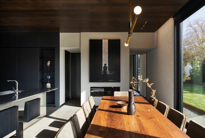 Modern dining room with a wooden table, chairs, and a minimalist black kitchen. Large windows provide natural light. Artwork on wall, vase with branches on table. Greenery visible outside.