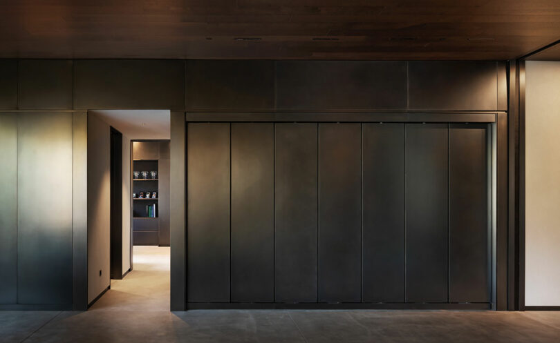 Minimalist interior with metal sliding doors closed on the right and a doorway open to a dimly lit room with shelves on the left. Walls and ceiling have a dark finish.