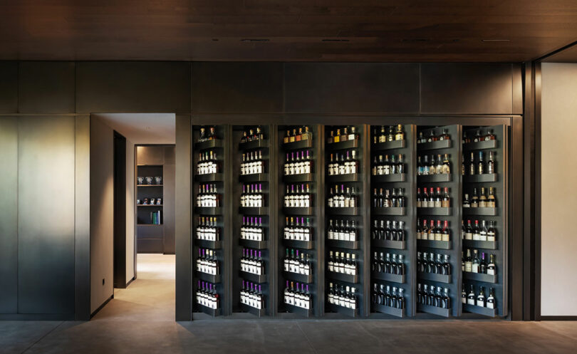 A wall of neatly arranged wine bottles in a dimly lit room with additional storage visible through an open doorway.