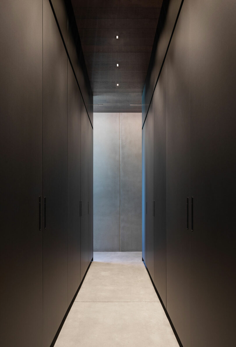 Minimalist hallway with dark walls and overhead lights leading to a softly lit doorway.