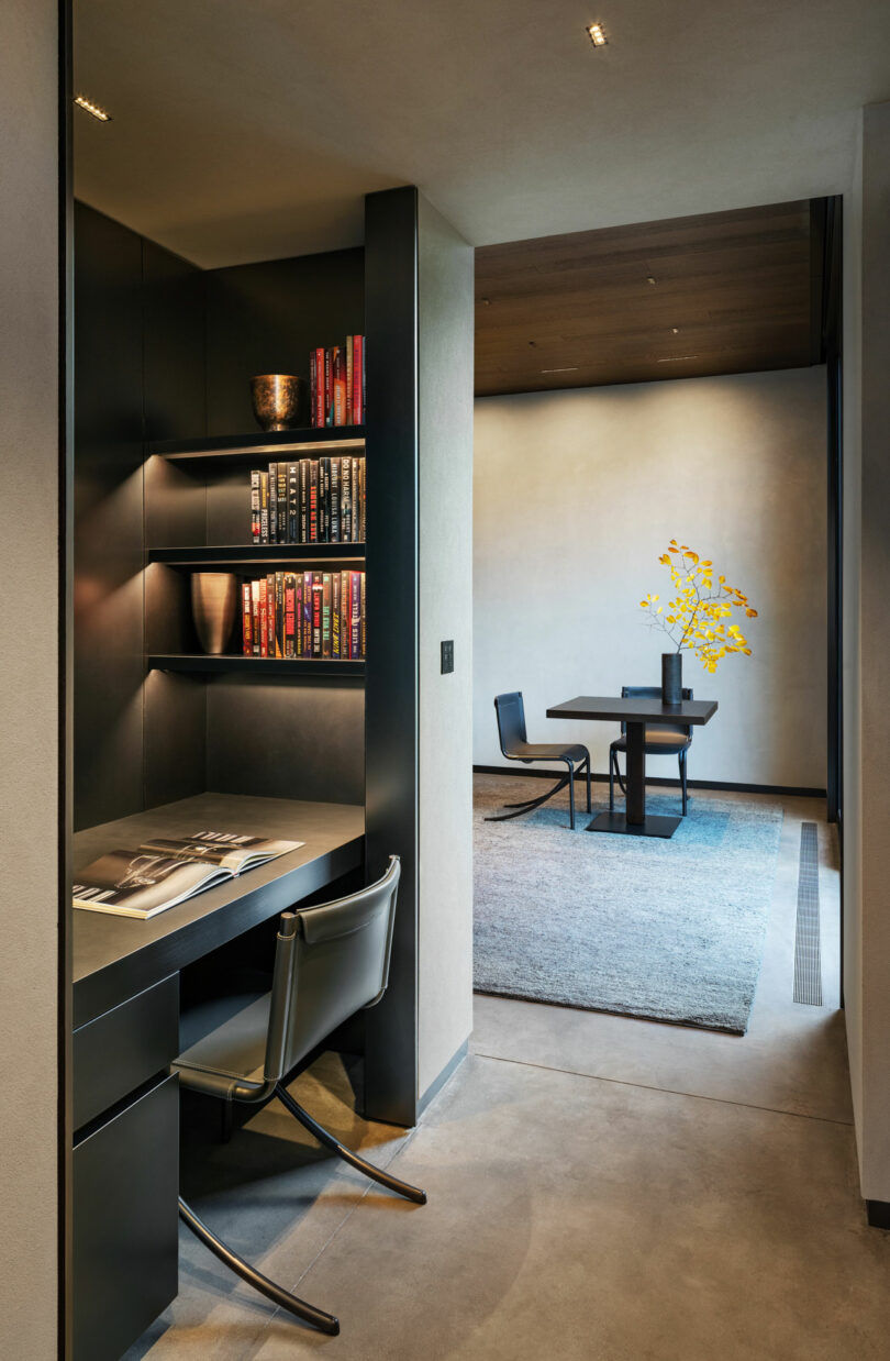 Interior moderno con un rincón de estudio con un escritorio, silla y estantería. En el fondo, una mesa de comedor con sillas y un jarrón con flores amarillas en una alfombra.