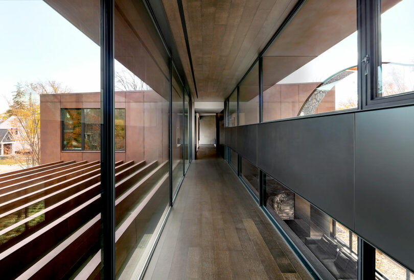 Modern architectural interior with a narrow corridor featuring glass walls and a wooden floor, connecting two sections of a building. Horizontal beams are visible through the glass.