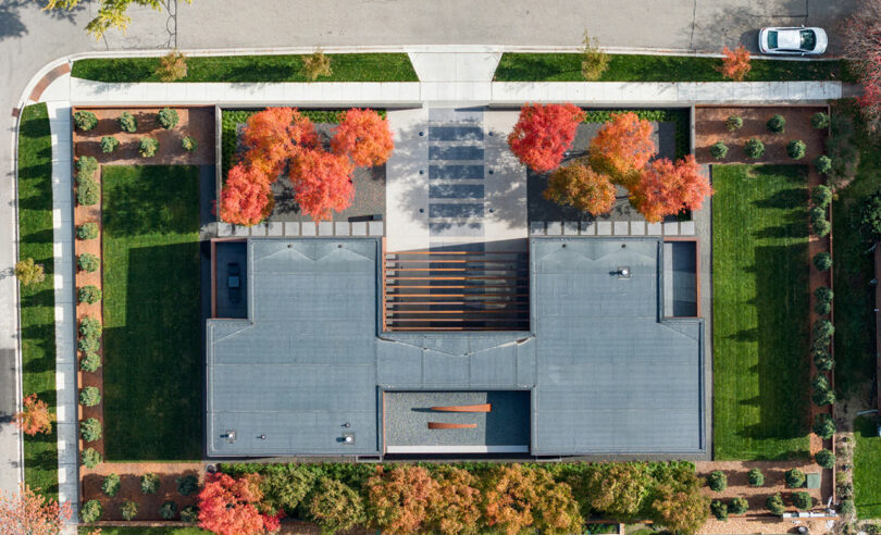 Aerial view of a modern building with a flat roof, surrounded by landscaped lawns and autumn trees. A car is parked on the street above the property.