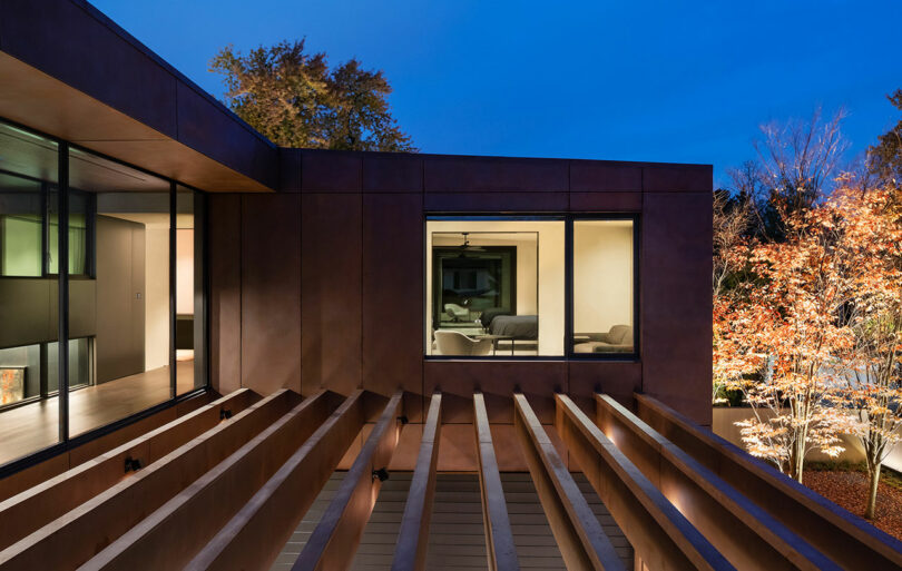 Modern house exterior at dusk, featuring large windows and wooden beams. Warm lighting inside contrasts with the blue evening sky. Sparse trees with autumn leaves are visible in the courtyard.