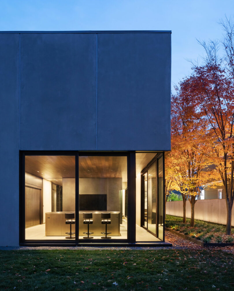 Edificio moderno con grandes vidrio que revela un interior brillante con sillas y una mesa. Está adyacente a un césped y un árbol con hojas de color otoño, ubicado contra un cielo crepuscular.