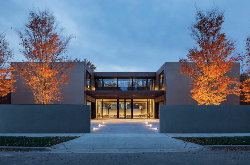 Modern two-story house with large glass windows, flat roof, and illuminated autumn trees on both sides, viewed from the street.