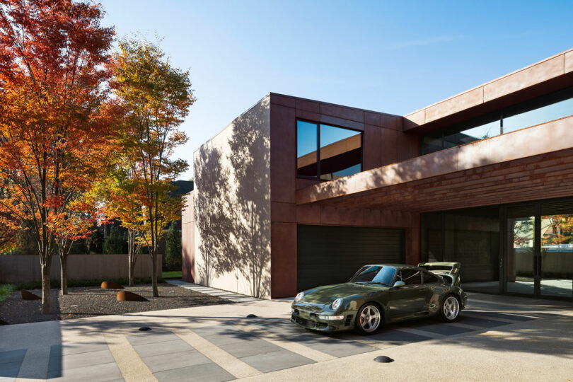 Casa moderna con ventanas grandes y una fachada marrón, con un automóvil deportivo verde oliva estacionado en el camino de entrada. Los árboles de otoño con hojas naranjas están a la izquierda.