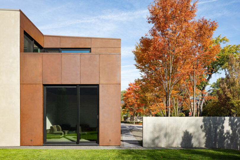 Exterior moderno de la casa con grandes ventanas de vidrio, rodeadas por un césped verde y vibrantes árboles de otoño.