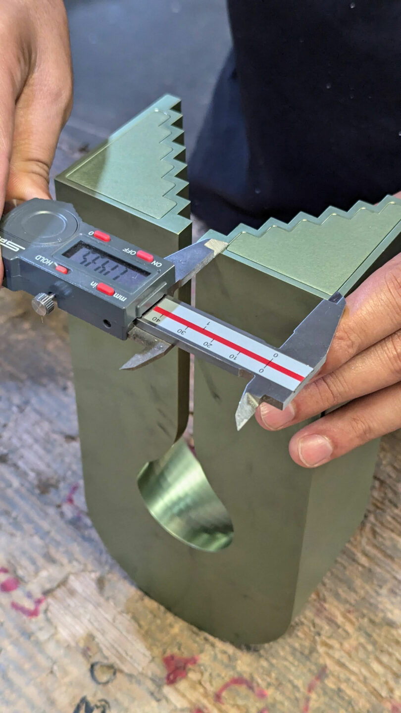 Hands using a digital caliper to measure the width of a green metal object with a U-shaped cutout.