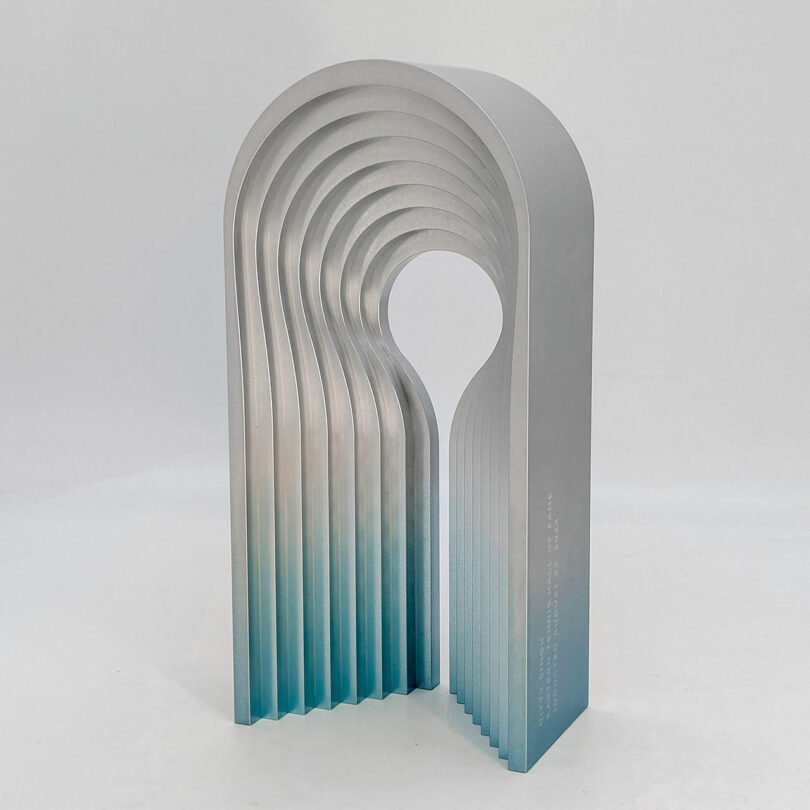 A metallic, arch-shaped sculpture with layered, gradient blue to silver tones on a plain white background.