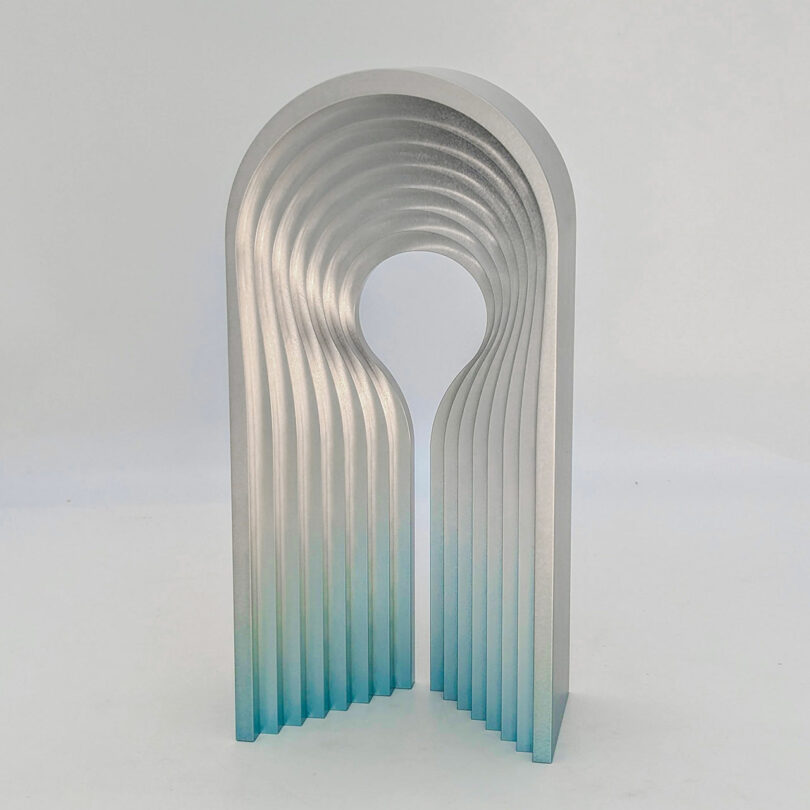 A silver metallic sculpture with a gradient blue base, featuring an arch shape with parallel curved ridges and a circular cutout in the center.