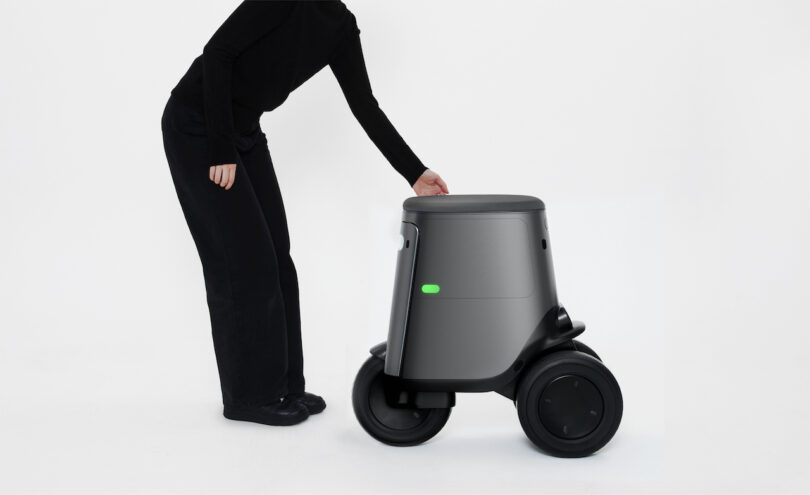 A person in black clothing interacts with a small, gray, wheeled robot on a white background