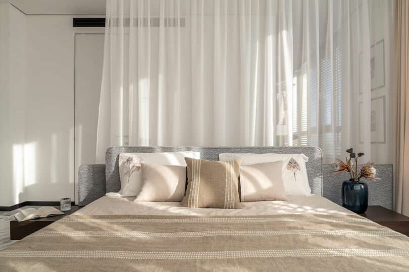 Modern bedroom with a neatly made bed, light beige bedding, and decorative pillows. Sheer curtains hang above, with sunlight streaming in. A book and vase with dried flowers are on the bedside tables.