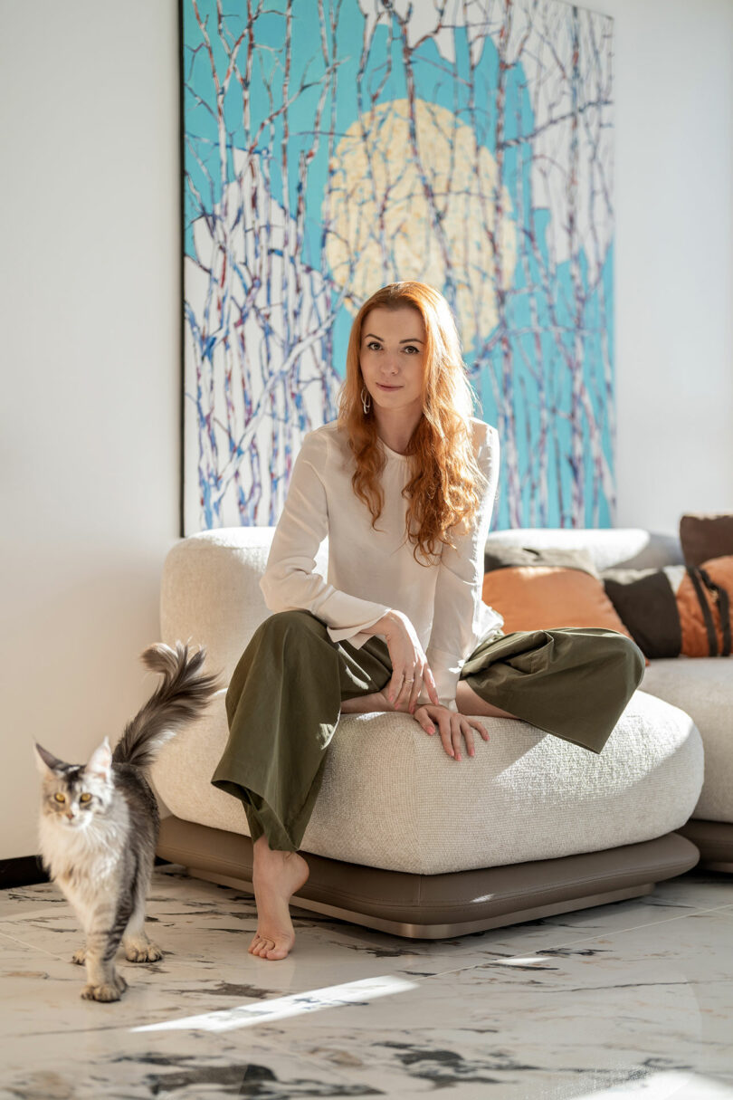 Woman with long red hair sitting on a white couch, wearing a white top and green pants, next to a gray cat. Behind her, a large abstract painting with blue and beige tones is on the wall.