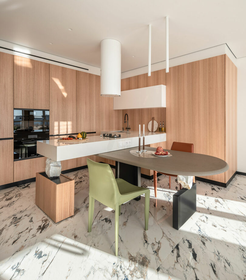 Modern kitchen with wooden cabinets, marble floor, an oval dining table, and mismatched chairs. A minimalistic design with a countertop featuring a built-in stove and a range hood above.