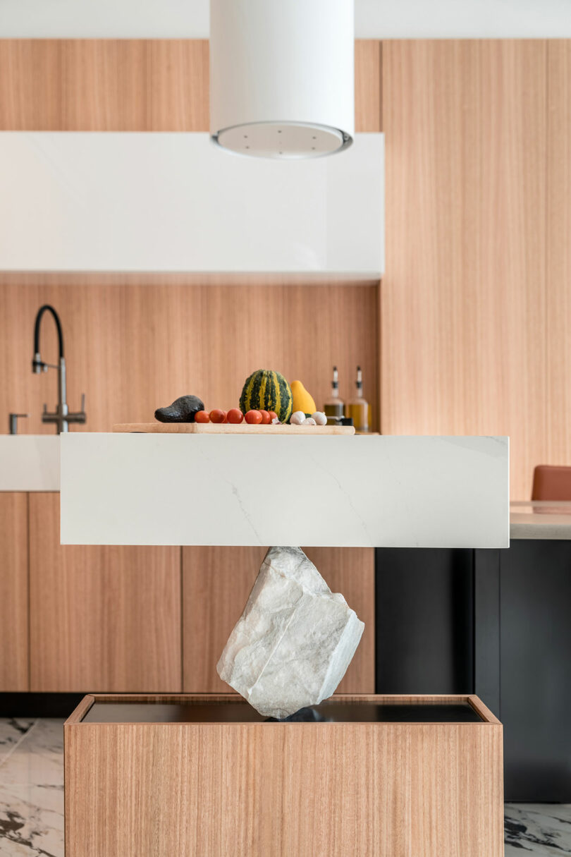 Modern kitchen with wooden cabinets, a sink, and a floating countertop. A balanced stone supports the countertop, with assorted fruits and vegetables on top. Marble floor visible.