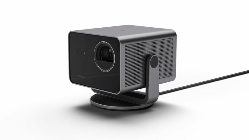 The Leica Cine Play 1 is a sleek, black rectangular projector featuring a circular lens and perforated side on a stand, equipped with a power cable extending to the side.