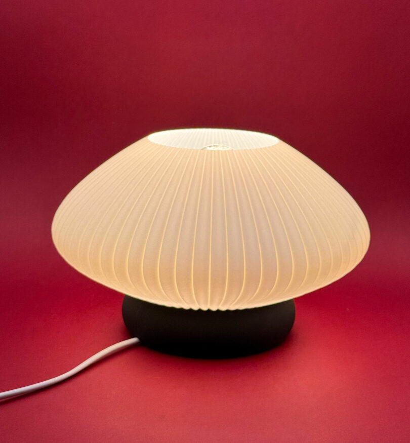 A mushroom-shaped table lamp with a pleated white shade emits a soft glow against a red background. A white power cord is visible.