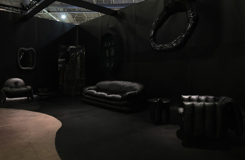 A dimly lit room at Maison&Objet showcases dark, cushioned furniture—sofa, chairs, and small tables—against black walls. Curved design elements intertwine gracefully with shadows, creating an atmosphere of intrigue and elegance.