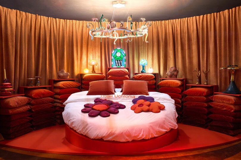 A Maison&Objet-inspired circular bed features colorful flower-shaped pillows, surrounded by curtain-draped walls and warm lighting. A decorative chandelier and stained glass window add an elegant touch to the room.