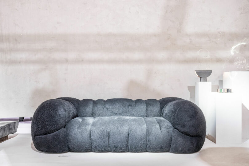 A plush gray sofa with rounded armrests graces this Maison&Objet-inspired minimalist room, featuring white walls and scattered decorative objects.