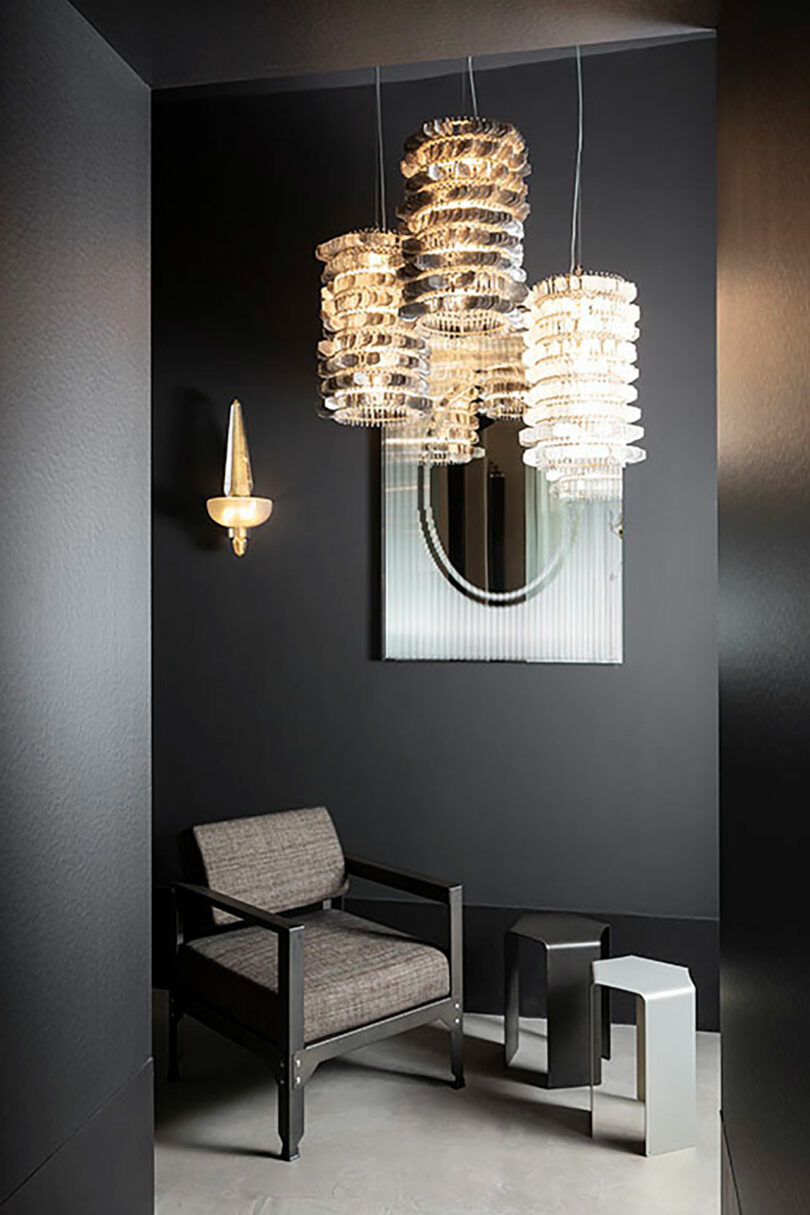 A modern interior showcases a Maison&Objet gray armchair, two metallic side tables, an elegant wall mirror, and three stacked glass pendant lights against dark walls.