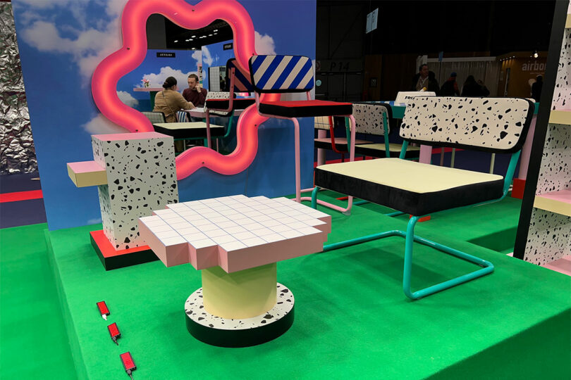 At Maison&Objet, discover a colorful, geometric furniture display featuring a grid-patterned table and abstract chairs on a green floor. Neon lights illuminate patterned surfaces, while cloud wallpaper adds depth to the vibrant scene.