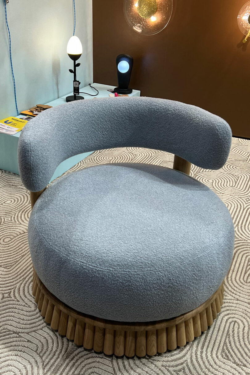 A round, plush chair with light blue fabric upholstery and a curved backrest on a wooden base graces a textured, patterned floor. Inspired by Maison&Objet, decorative lights and a table create an elegant backdrop.