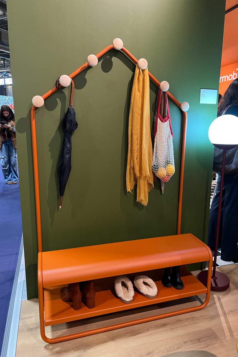 Decorated with Maison&Objet flair, the orange metal coat rack with a bench sits stylishly against a green wall. It holds an umbrella, scarves, and a net bag, while shoes neatly rest on the lower shelf as someone enters through the door on the left.