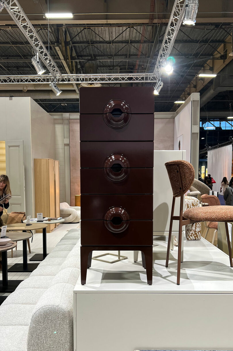 In the Maison&Objet showroom, a tall, dark brown drawer cabinet with circular handles stands elegantly on a platform, surrounded by an array of exquisite furniture pieces.