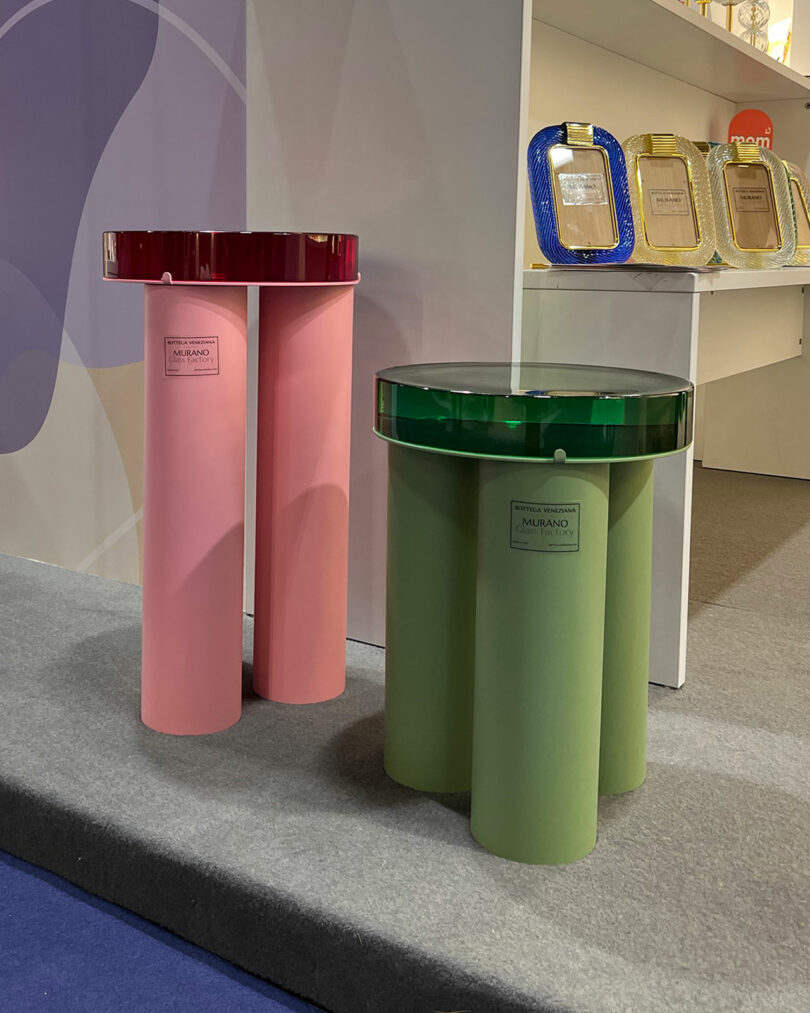 Two cylindrical tables with glass tops, one pink with a red top and one green with a green top, elegantly showcased at Maison&Objet on a carpeted floor, surrounded by decorative items on shelves behind.