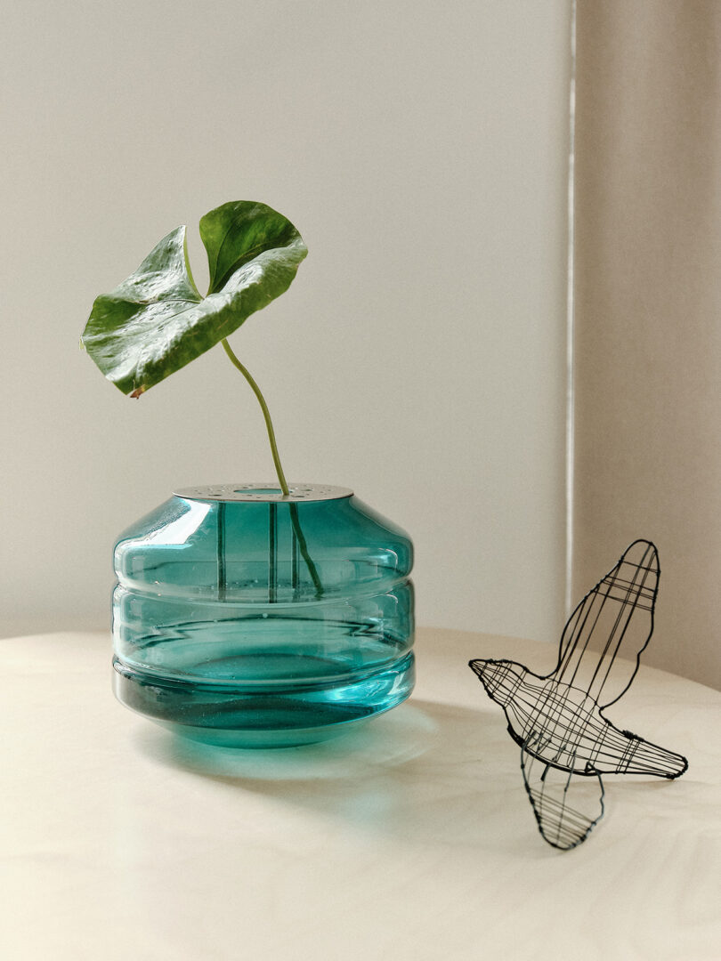 A single green leaf in a translucent blue vase on a light surface, next to a wire sculpture of a bird.