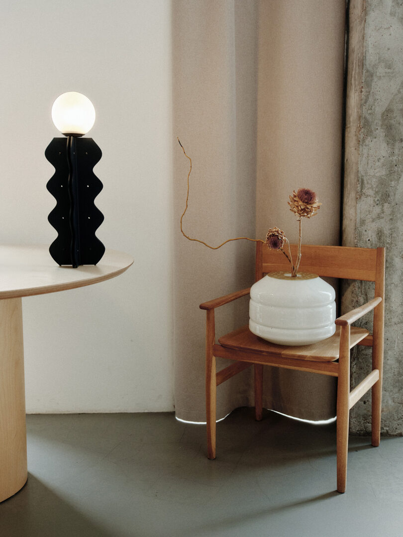 A wooden chair with a large white vase holding dried flowers sits beside a round table with a black zigzag lamp, against a beige curtain.