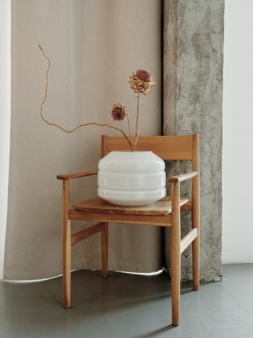 A wooden chair with a white vase holding dried flowers sits beside a concrete column on a gray floor, partially backed by a beige curtain.