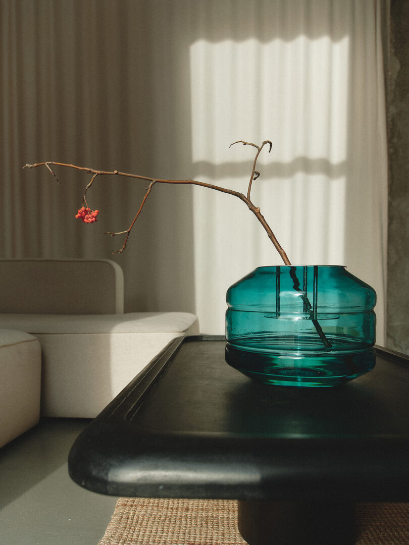 A branch with red berries in a teal glass vase on a black table. A beige sofa and curtain are visible in the background. Natural light casts shadows.