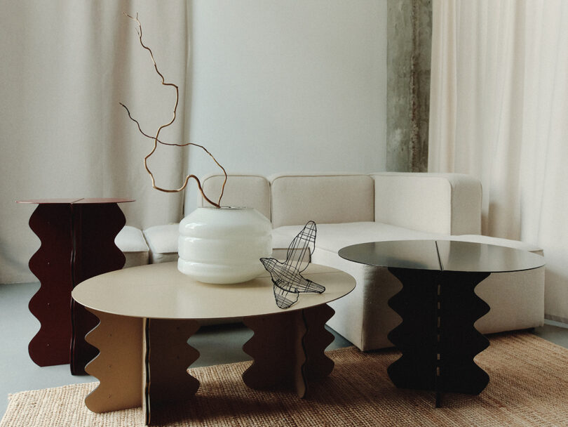 Modern living room with a beige sectional sofa, three wavy-edged tables, a white vase with branches, and a wire butterfly sculpture on a woven rug.