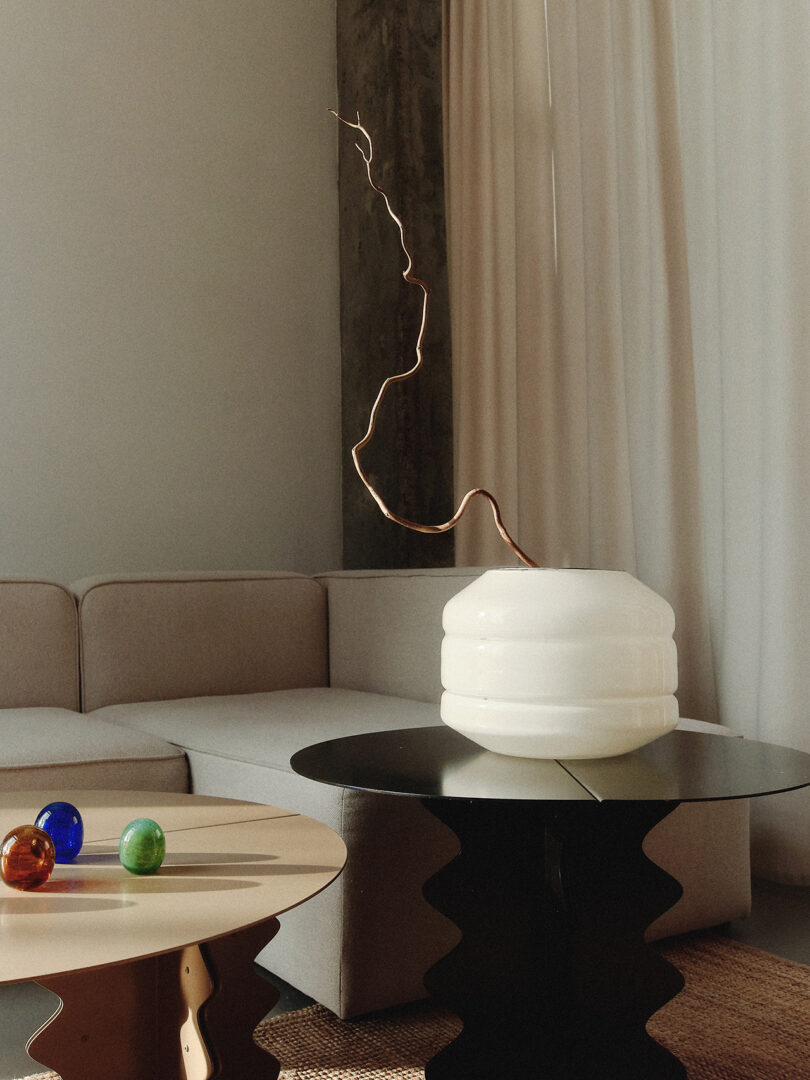 Minimalist living room with a beige sectional sofa. A modern white vase with a branch sits on a black table. Colorful decorative spheres are on a light wooden table. Light curtains in background.