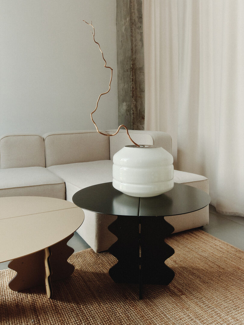 A minimalist living room with a beige sofa, two artistic black tables, a woven rug, and a white vase with a twisted branch.